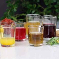 several glasses filled with different colored liquids on top of a white countertop next to vegetables and flowers