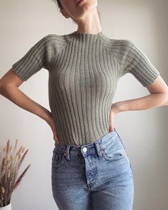 a woman standing in front of a white wall wearing jeans and a gray knitted sweater