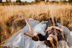 two people laying on a blanket in the middle of a field with tall grass and trees