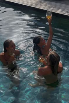 three women in a pool drinking wine and toasting with their hands up to the sky