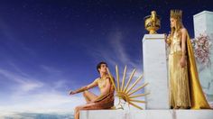 two women dressed in gold sitting next to each other on top of a pedestal with a sun