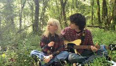 two people sitting in the woods playing guitars