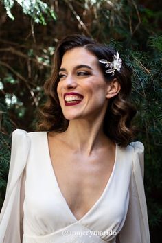 a woman in a white dress smiling and wearing a flower on her hair with greenery behind her