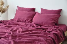 an unmade bed with purple sheets and pillows on it, next to a plant