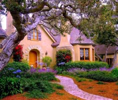 a yellow house surrounded by lush green trees and purple flowers in the front yard, with a pathway leading to it