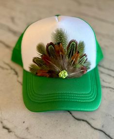 This beautiful green & white trucker hat with green and natural colored feathers, and a green stone embellishment will be all anyone can talk about when you wear it.  It's such a fun way to spice up any outfit and the places to wear this hat are endless. Be prepared for loads of compliments.  The feathers are handpicked for each hat.  The hat has a foam front, mesh back, and an adjustable snap back. Adjustable Green Costume Hats With Short Brim, Adjustable Green Hats For Festivals, Adjustable Green Hat For Festival, Adjustable Green Hat For Festivals, Green Short Brim Hat For Festivals, Green Festival Cap, Adjustable Brimmed Green Mini Hats, Adjustable Green Brimmed Mini Hats, Green Trucker Hat For Spring, One Size Fits Most