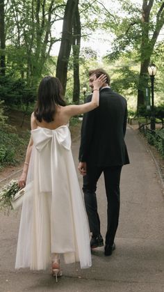 a bride and groom walking down a path in the woods together, holding each other's hands