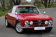 an old red alfa car is parked on the side of the road in front of some trees