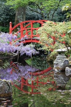 a red bridge over water surrounded by rocks and trees with purple flowers in the foreground