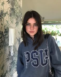 a woman wearing glasses and a sweatshirt with the word pst on it, standing in front of a wall