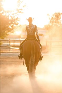 a woman riding on the back of a brown horse across a dirt field at sunset