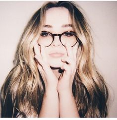 a woman with long hair and glasses posing for the camera in front of a white wall