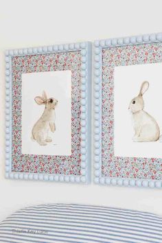 two framed pictures of rabbits on a white wall above a blue and white striped bed