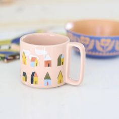 a pink coffee mug with houses painted on it next to a bowl and pencils