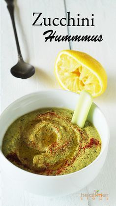 a white bowl filled with green dip next to a lemon slice and spoon on a white table