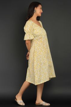 a woman in a yellow and white dress standing against a gray background with her hands on her hips