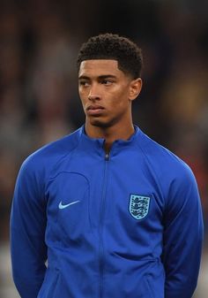 soccer player in blue jacket standing on field with head turned to the side and looking at camera