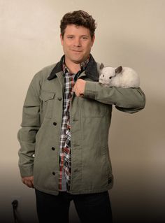 a man holding a small white animal on his arm while standing in front of a wall