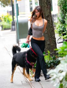 a woman walking her dog on a leash down the sidewalk in front of a tree