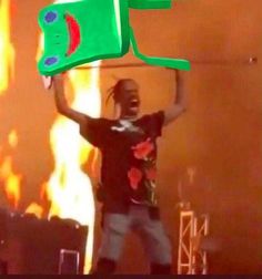 a man holding up a large green object in front of a fire and flames background