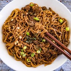 a white bowl filled with beef and noodles next to chopsticks on a wooden table