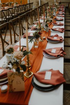 a long table set with place settings and orange napkins on it, along with candles