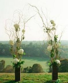 two tall vases with flowers in them on top of a grass covered hill next to trees