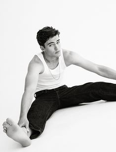 a young man sitting on the floor with his legs spread out, wearing jeans and a tank top