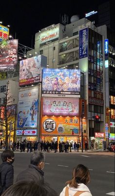 many people are standing on the street in front of tall buildings at night with signs and advertisements