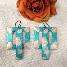 two square shaped blue and pink earrings next to a flower on a white cloth with an orange rose in the background