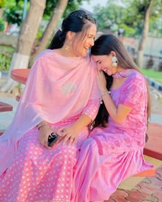 two women in pink dresses sitting on a bench and hugging each other with trees in the background