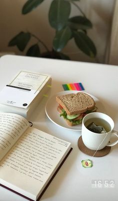 an open book, coffee cup and sandwich on a white table with a plant in the background