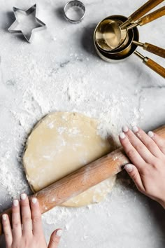 two hands rolling dough on top of a table next to baking utensils and cookie cutters