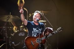 a man holding a guitar and giving the peace sign while standing in front of drums