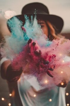 a woman wearing a hat and holding her hands up with colored powder in the air