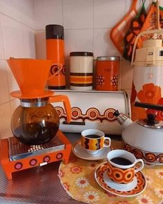 an orange and white coffee pot sitting on top of a wooden table next to two cups