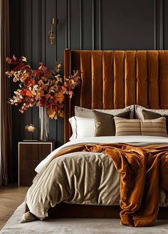 a bed with brown sheets and pillows next to a tall vase filled with autumn leaves