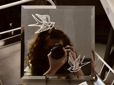 a woman taking a photo with her camera in front of a mirror that has birds drawn on it