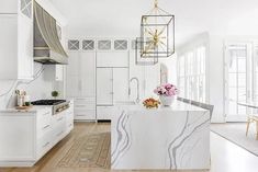 a white kitchen with marble counter tops and gold pendant lights hanging from the ceiling over the island