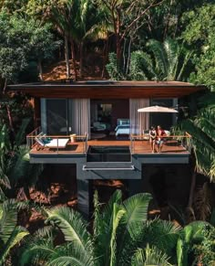 two people sitting on the deck of a house surrounded by palm trees and greenery