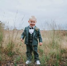 a young boy in a green suit and bow tie standing in tall grass smiling at the camera