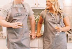 two people in aprons holding drinks and smiling at each other while standing in the kitchen