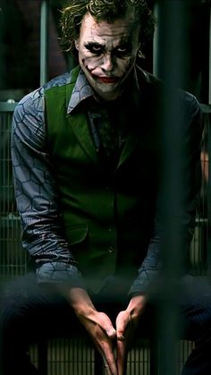 the joker is sitting in his caged area with his hands on his chest and looking at the camera