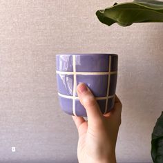 a hand holding a purple coffee cup with white lines on the outside, and a green plant behind it