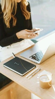 a woman sitting at a table looking at her cell phone and laptop with a cup of coffee in front of her