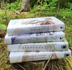 three books are stacked on top of each other in the grass near some trees and bushes