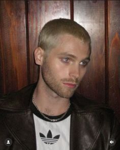 a man in a leather jacket and adidas t - shirt looking at the camera