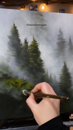 a person holding a pen in front of a painting with trees on it and fog