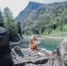 a woman sitting on the edge of a cliff looking at a map in front of a mountain lake