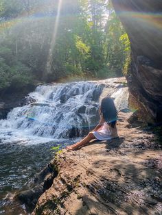 a woman sitting on top of a rock next to a waterfall in the forest with sun shining down